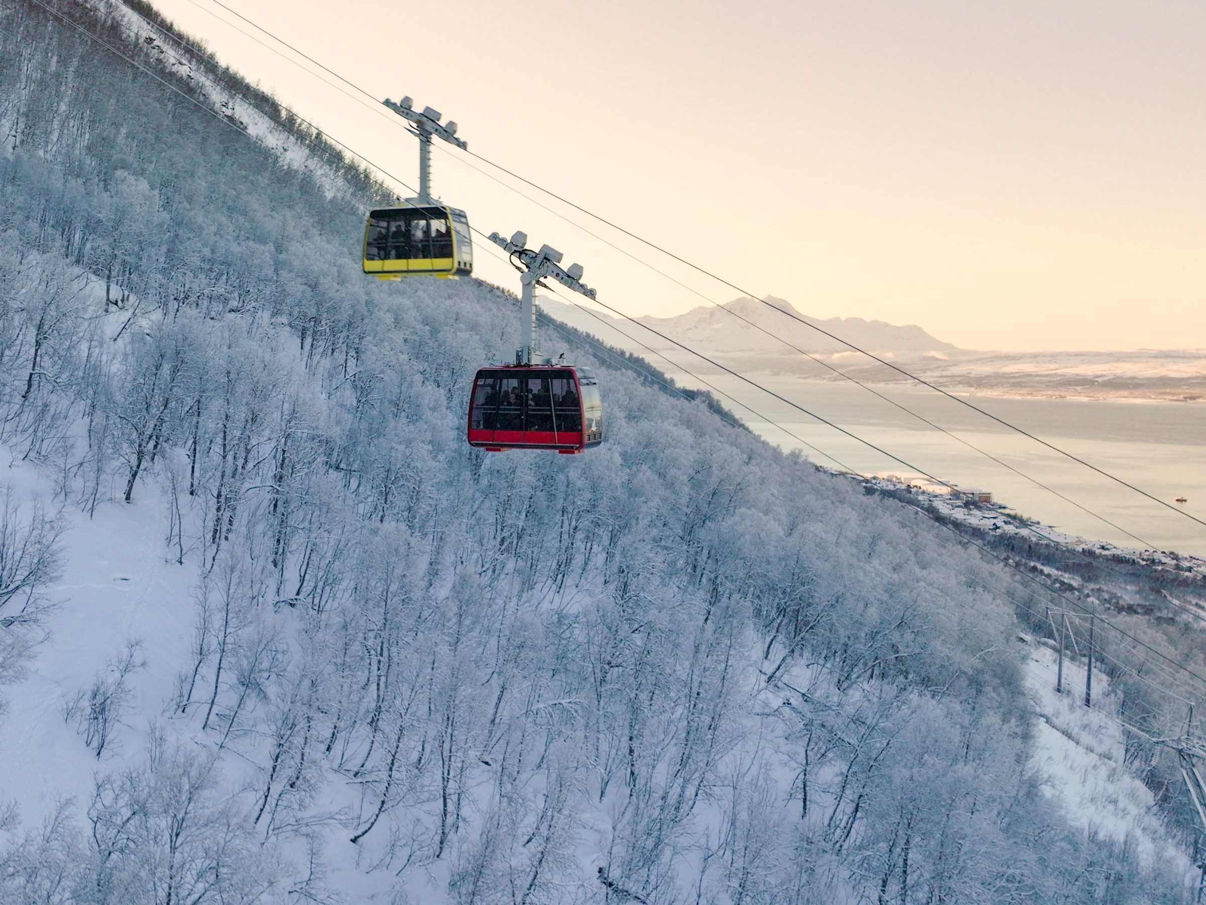Two cable cars on snowy mountain with a view of a fjord.
