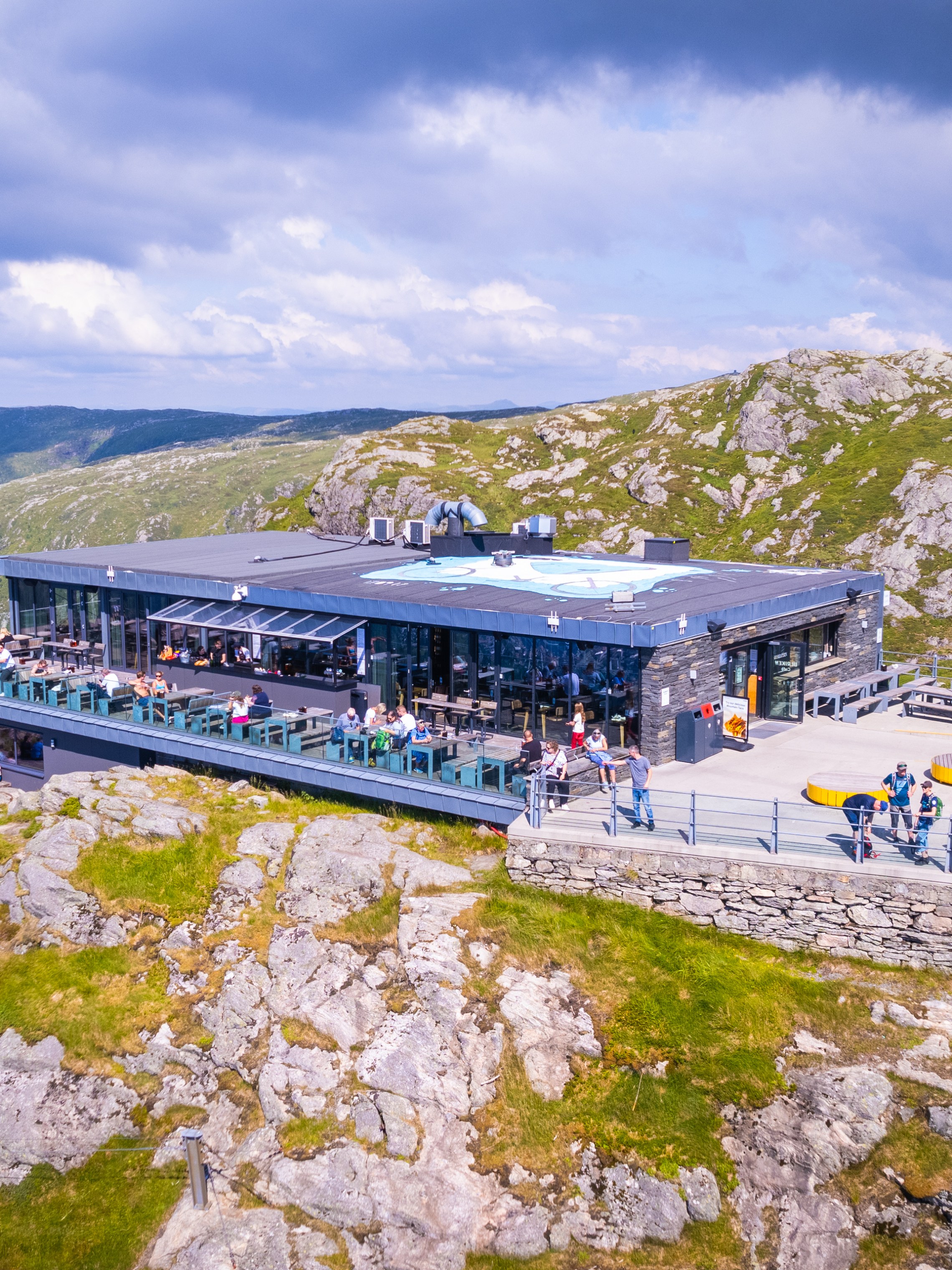 Mountain restaurant with outdoor seating, Norwegian flag, overlooking scenic rocky landscape.