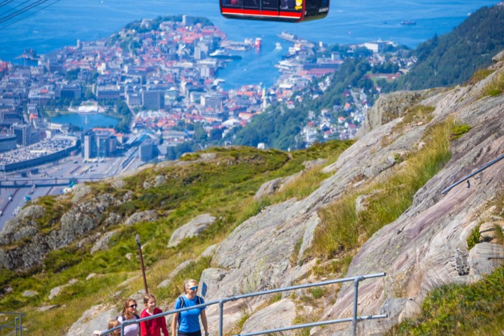 Three hikers on a rocky path beside a cable car with a city and fjord view in the background.