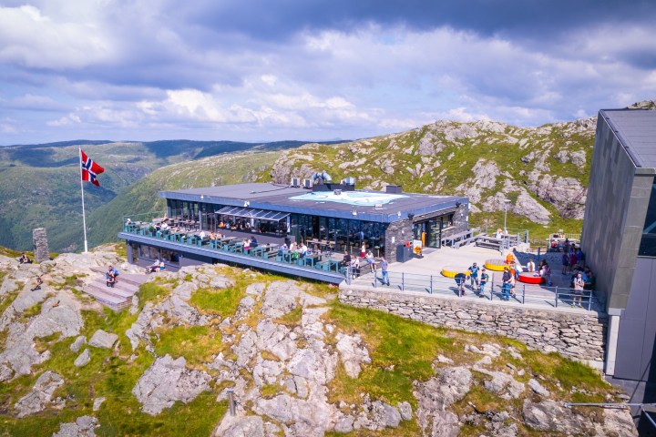Hilltop building with a Norwegian flag surrounded by rocky landscape and tourists under a cloudy sky.