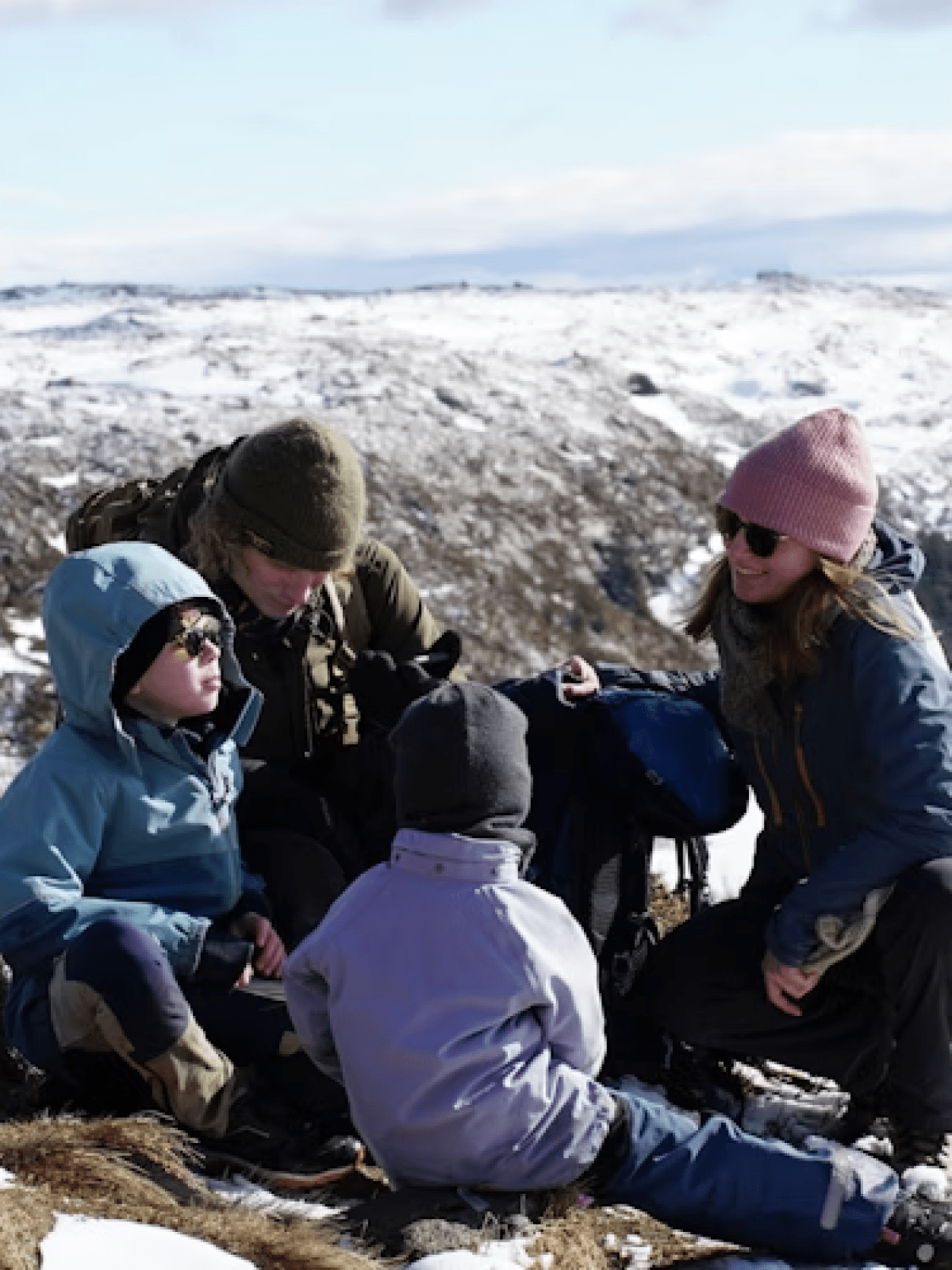 Family sitting on snowy hillside with backpacks, wearing winter clothing.