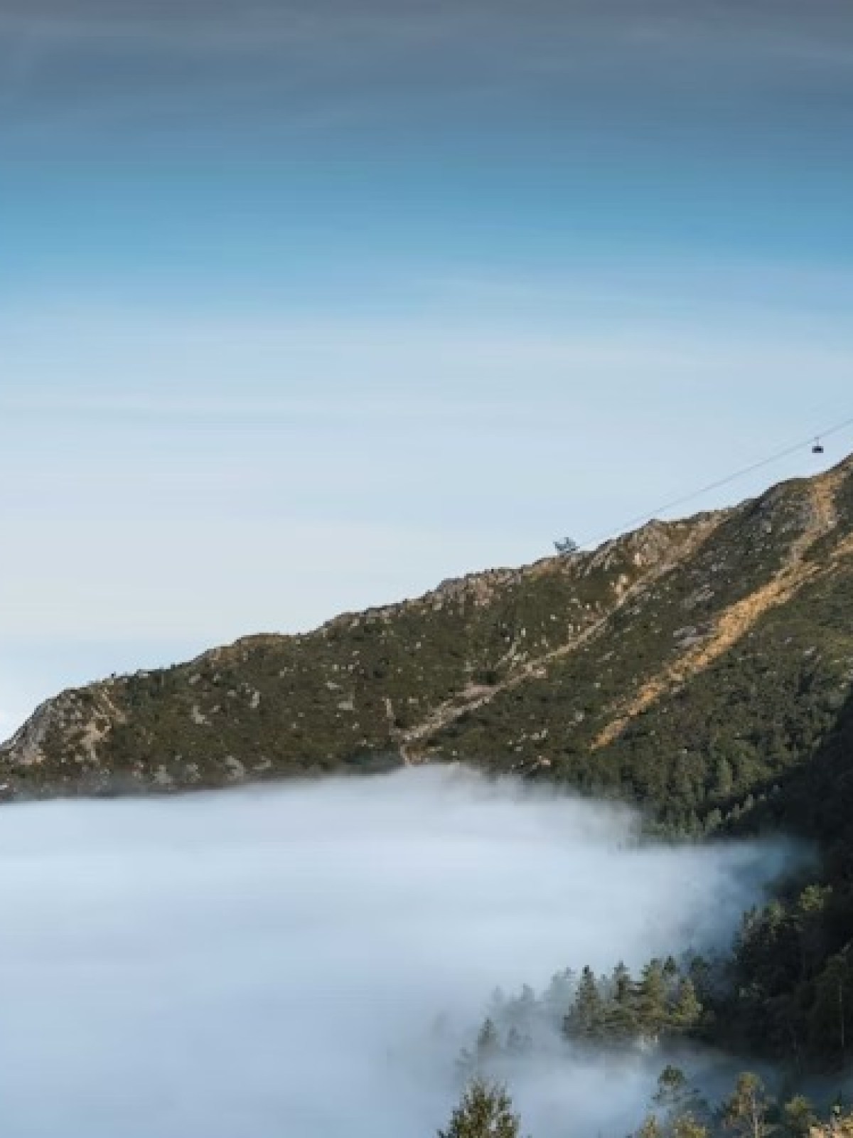 Mountain peak with a cable car above clouds under a clear blue sky.