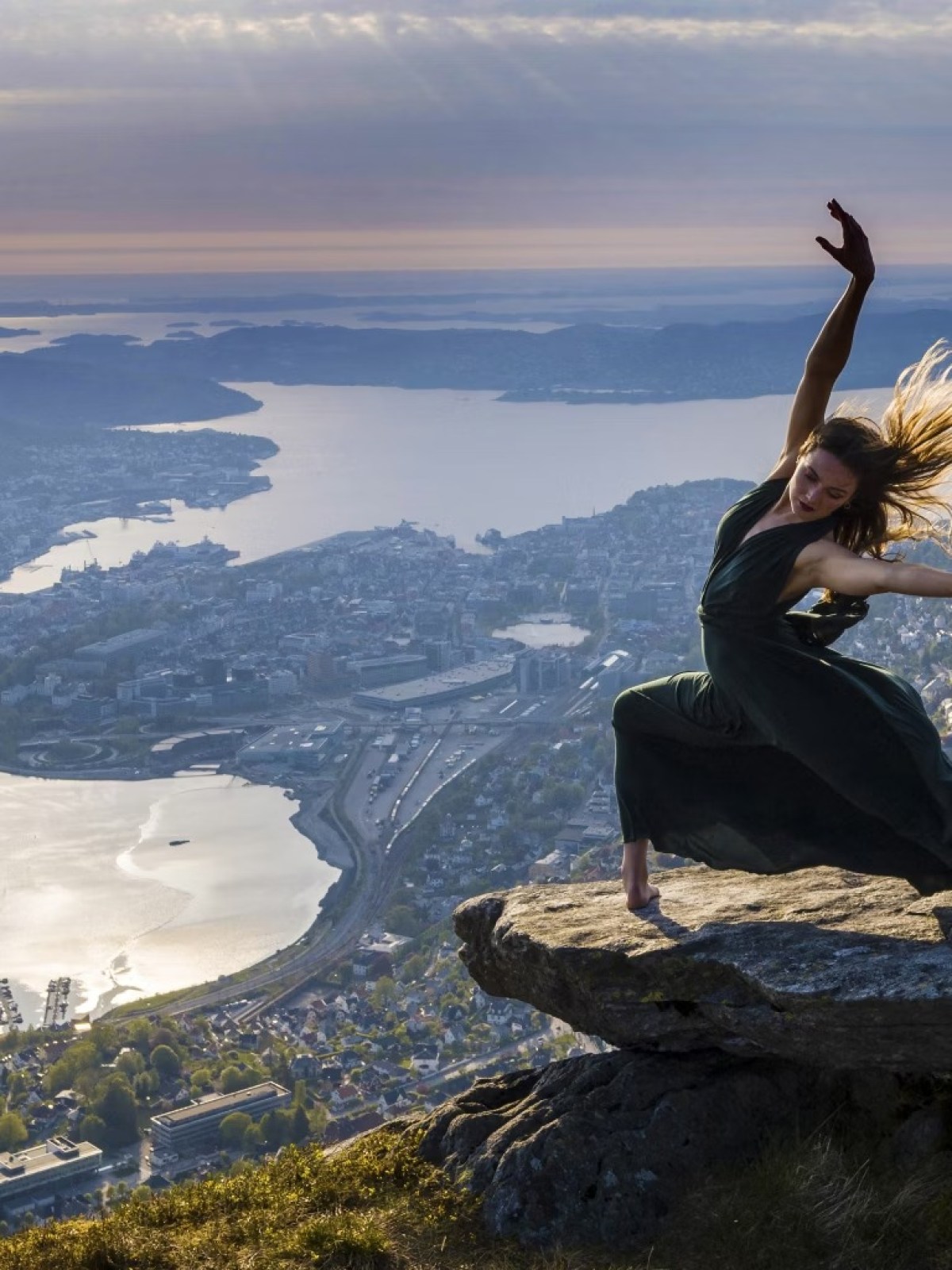 Woman in a flowing dress dances on a rocky ledge above a cityscape at sunset.