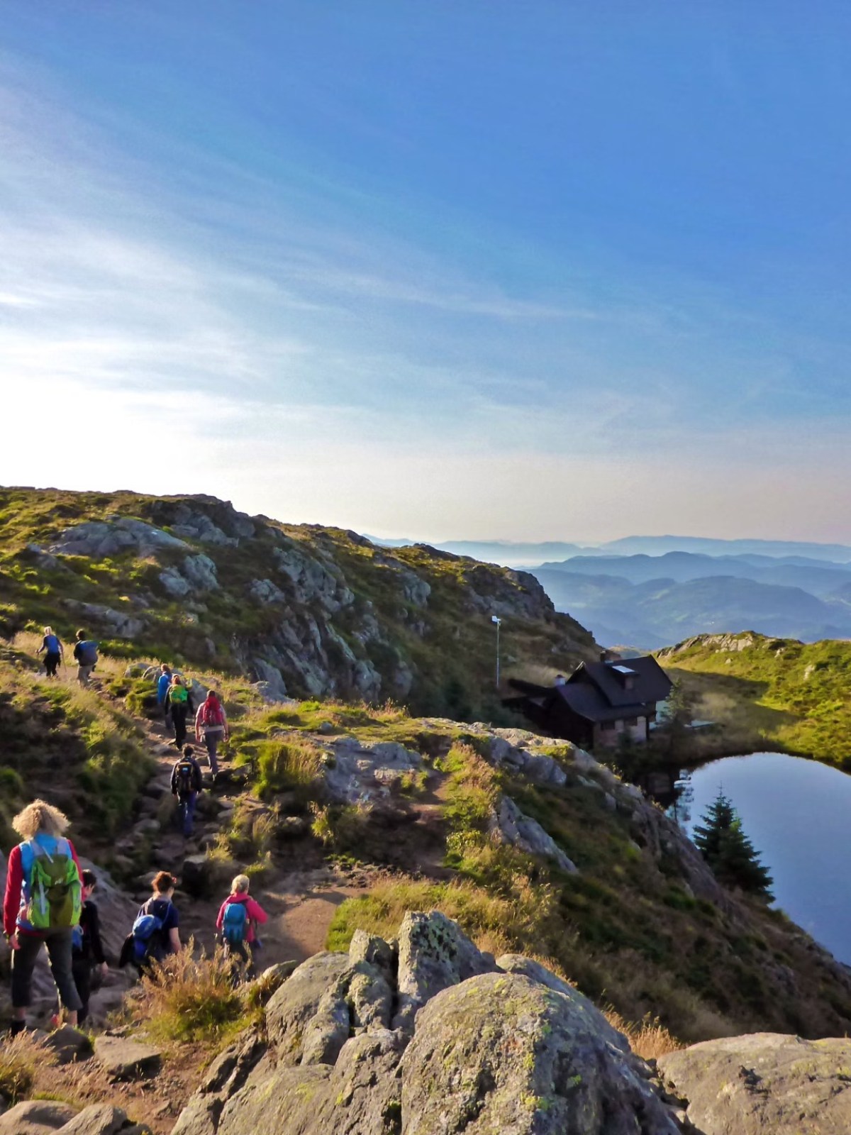Group hiking on mountain trail near a pond under a clear blue sky.