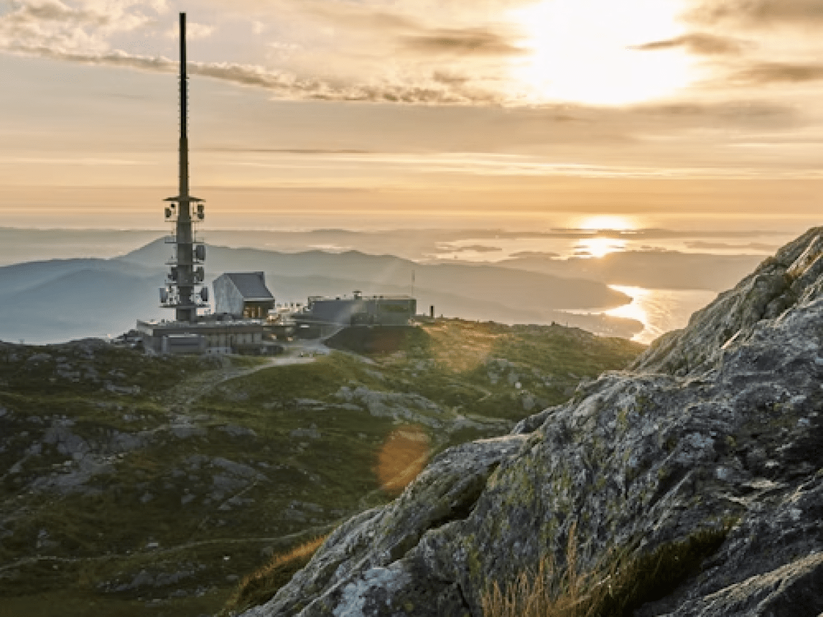 Antenna tower on a mountain at sunset with rocky foreground and distant hills.